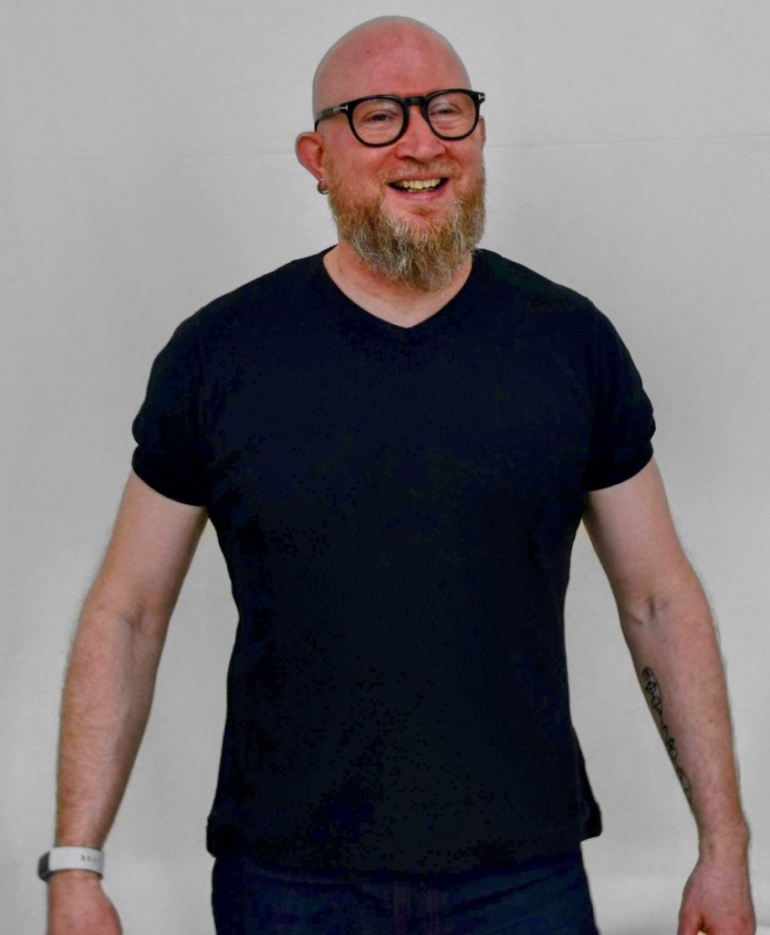 bald man with thick glasses in t-shirt smiling at someone behind the camera